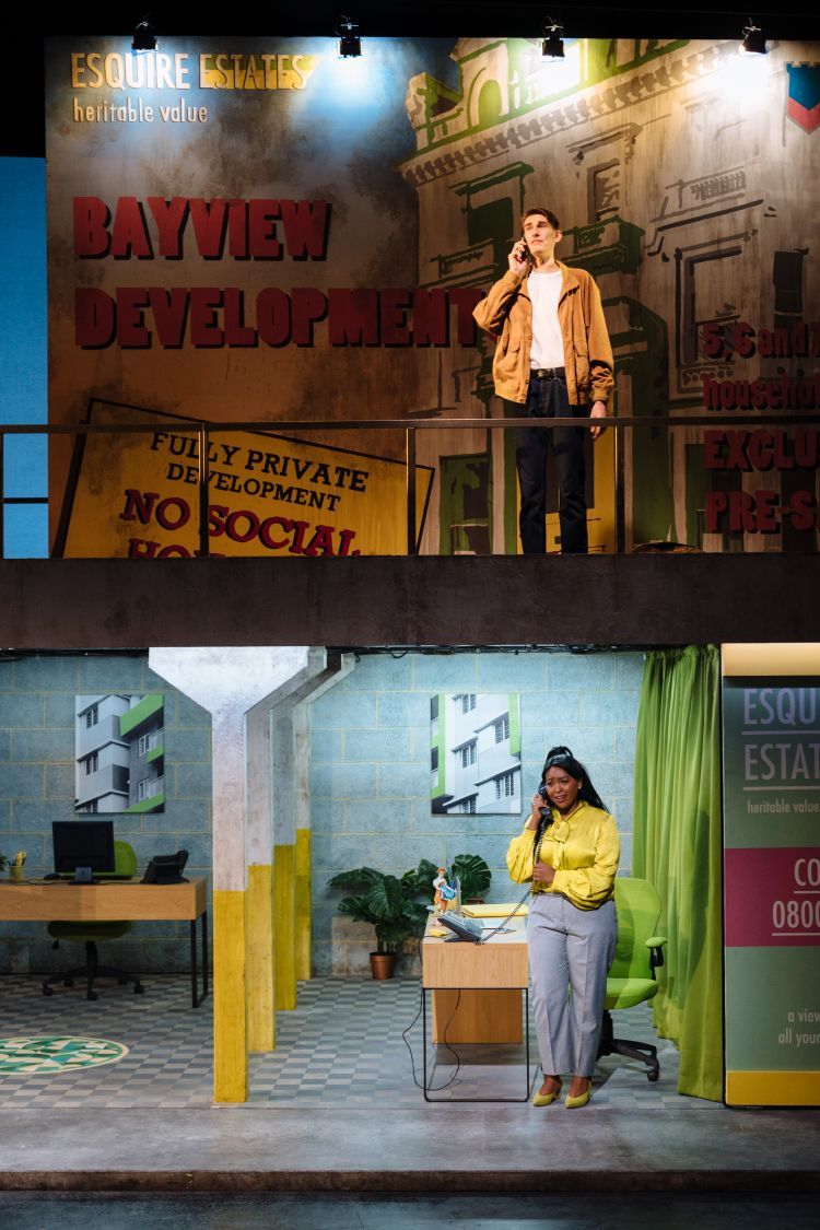 Two singers speak into telephones on a split-level stage, one standing a level higher than the other